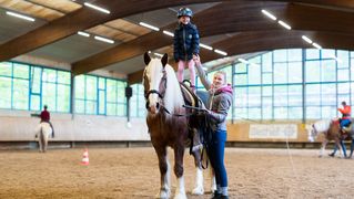 Ein Mädchen steht auf einem Pferd. Eine junge Frau steht neben dem Pferd und hält die Hand des Mädchens.