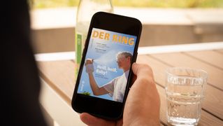 Eine Person sitzt an einem Tisch mit einem Glas Wasser und hält ein Smartphone mit der neuesten Ausgabe von DER RING in der Hand.