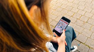 Eine Frau sitzt auf einer Mauer und hält in der Hand ein Smartphone mit dem Cover von DER RING