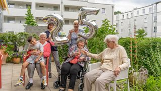 Frauen sitzen und stehen auf einer Terasse. Die älteren Bewohnerinnen stoßen mit Sekt an und die Mitarbeiterinnen halten Luftballons mit der Zahl 25 hoch.