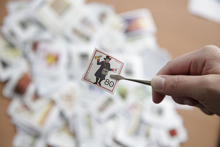 A stamp is held up to the camera with a pair of tweezers. In the background, many more stamps are lying on a table.