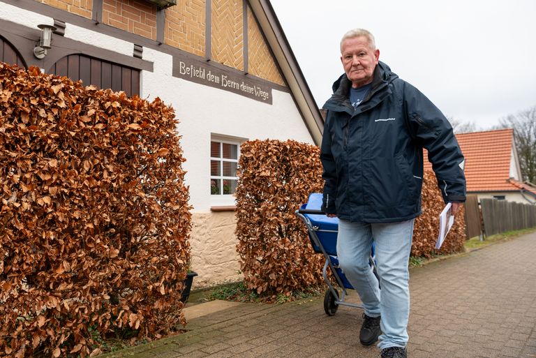 Andreas Zylla geht auf dem Bürgersteig an einem Haus vorbei. Er zieht einen Rollwagen hinter sich her. In der linken Hand hält er eine Zeitschrift.