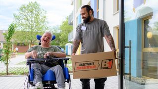 Zwei Männer, einer davon nutzt einen Rollstuhl, kommen in ein Gebäude.